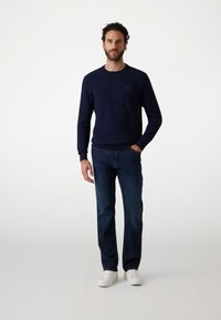 Felpa leggera blu navy con colletto rotondo e polsini a costine, abbinata a jeans scuri in denim e sneakers bianche. Branding discreto incluso.