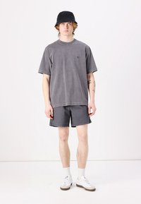 Šedé oversized tričko a odpovídající šortky, obě z jersey materiálu. Model má na sobě černý klobouk typu bucket a bílé tenisky se šedou podrážkou.