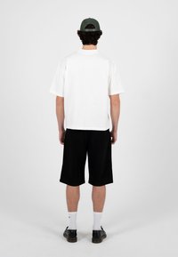 Mann som står med ryggen til, iført en hvit kortermet skjorte, svarte knebukser, hvite sokker, svarte sko og en grønn baseballcaps.