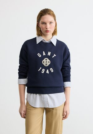 Ung kvinne med kort hår som har på seg en blå GANT 1949-genser over en stripete skjorte og beige bukser, stående foran en ensfarget hvit bakgrunn.