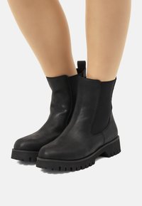 Botas de cuero negro hasta el tobillo con paneles laterales elásticos, punta redonda y suela de goma gruesa con un patrón de relieve y una pestaña en la parte posterior.