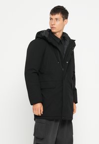 Zwarte gewatteerde parka met een capuchon, voorzien van knopen en een ritssluiting aan de voorkant, met twee zijzakken en een textuurstofafwerking.
