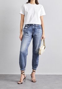 Witte cropped t-shirt, lichtblauwe denim joggers met versleten details, en zwarte bandjes hoge hakken. Houdt een kleine witte tas vast.