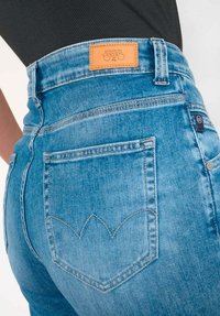 Des jeans en denim bleu taille haute présentent une étiquette en cuir, des coutures visibles et une poche arrière au design incurvé distinctif.