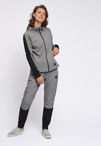 Haut de survêtement gris avec des accents noirs, fermeture éclair, et capuche ajustable ; associé à un pantalon de jogging gris et noir, tous deux en matière douce.