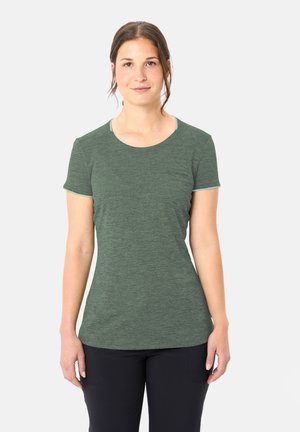 Frau mit zurückgebundenem braunem Haar, die ein eng anliegendes grünes Kurzarmshirt und schwarze Hose trägt und vor einem schlichten weißen Hintergrund steht.