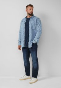 Lichtblauwe denim shirt over een marineblauw t-shirt, gecombineerd met donkere denim jeans en witte sneakers. Bevat een borstzak en een knoopsluiting aan de voorkant.