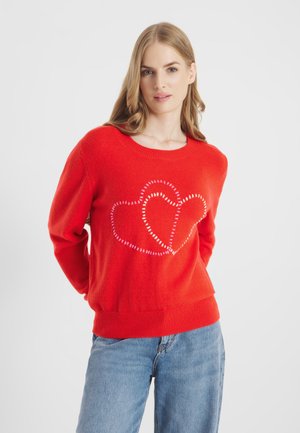 Kobieta w czerwonym swetrze z dwoma dużymi zszytymi sercami na przodzie, w jasnoniebieskich spodniach dżinsowych, stojąca na jednolitym białym tle.