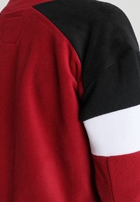 Punainen fleece-vaate musta-valkoisilla korostusunilla. Ominaisuuksina tekstuuri, pehmeä pinta ja takana paikka.