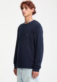 Mørkeblå strikket sweater med lange ærmer, rund hals og ribbede manchetter; har et lille logo på brystet; glat tekstur.