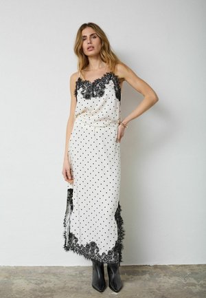 Woman in white slip dress with black polka dots, black lace trim, and black ankle boots, standing against a plain wall.