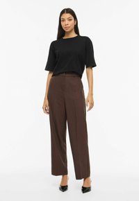 Mustast lühikeste varrukatega top koos kõrge vöökohaga pruunide laiade pükstega ja mustade terava varbaga kontsadega, taustaks lihtne valge sein.