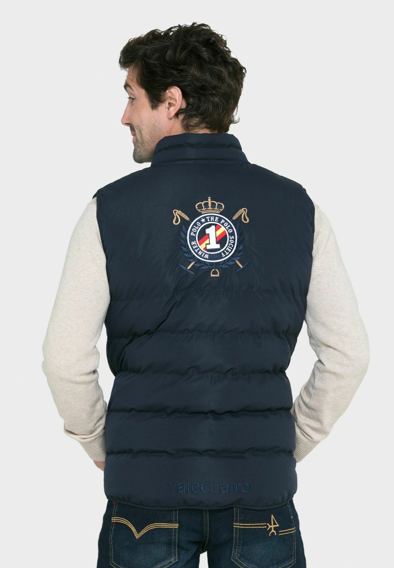 Chaleco acolchado azul marino con un diseño acolchado, que presenta un logotipo y bordado de corona en la parte posterior, combinado con una camiseta de manga larga gris.