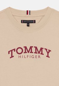 Bežinė medvilninė marškinė su apvaliu kaklu, su dideliu bordo spalvos "TOMMY HILFIGER" logotipu. Kaklaskare yra dryžuota detalė.