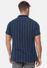 Koszula z krótkim rękawem i kołnierzykiem w ciemnym niebieskim kolorze z pionowymi czarnymi i białymi paskami. Teksturowana tkanina o luźnym kroju, szczegółowe przeszycia na kołnierzyku.