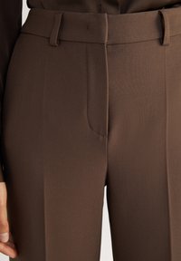 Gros plan sur un pantalon marron sur mesure avec passants de ceinture et fermeture avant dissimulée porté par une personne.