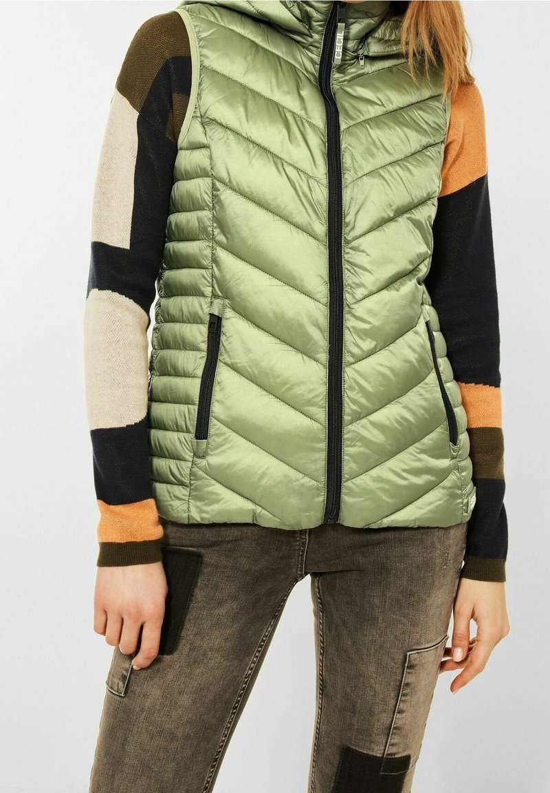 Personne portant un gilet sans manches matelassé vert clair par-dessus un pull patchwork multicolore à manches longues et un pantalon marron délavé avec des poches appliquées.