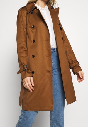 Frau trägt einen braunen doppelseitigen Trenchcoat mit schwarzen Knöpfen, weißes Hemd mit Kragen und blaue Jeans vor einem schlichten weißen Hintergrund.