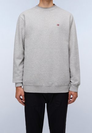 Homme portant un sweat-shirt gris clair uni à col rond avec un petit logo de drapeau rouge et blanc sur la poitrine gauche, assorti à un pantalon noir.
