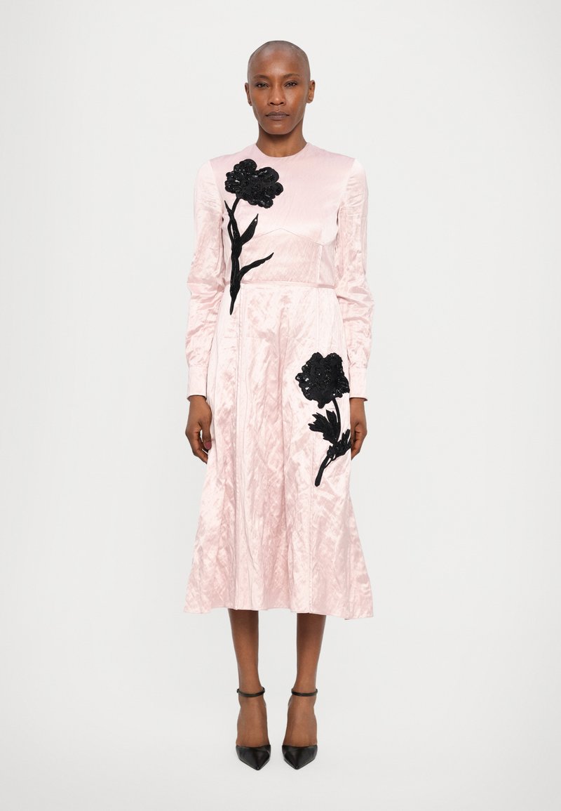 Long-sleeve pink dress with textured fabric, featuring black floral appliqués and a pleated skirt; ankle-strap black heels.