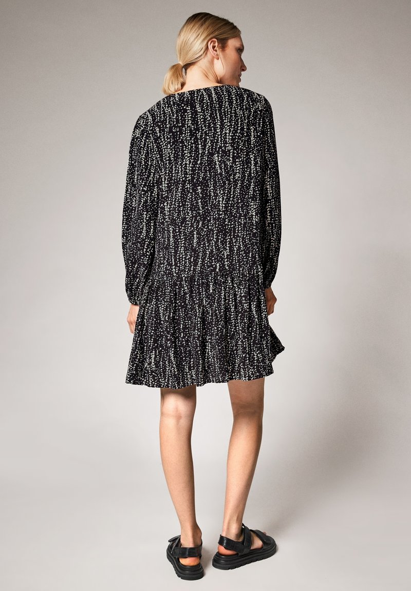 Kurzes schwarzes Kleid mit einem strukturierten Muster aus weißen Punkten, langen Ärmeln und einem ausgestellten Saum. Getragen mit klobigen schwarzen Sandalen.