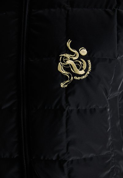 Sort pufferjakke med quiltet tekstur. Har et guld broderet logo på brystet. Frontlås med lynlås og en blød, glansfuld finish.