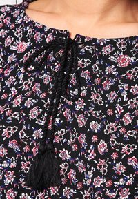 Blusa negra con flores que presenta un escote ajustable con cintas. La tela tiene una textura suave y muestra pequeños patrones en rojo, blanco y azul.