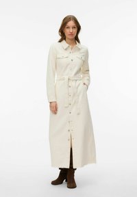 Femme portant une longue robe crème boutonnée avec ceinture et manches longues, associée à des bottes en daim marron, debout devant un fond blanc.