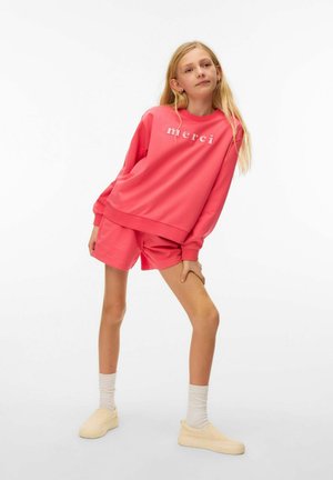 Blond meisje met een koraalroze sweatshirt met de tekst "merci", bijpassende korte broek, witte sokken en beige instappers, staand met één been gebogen.