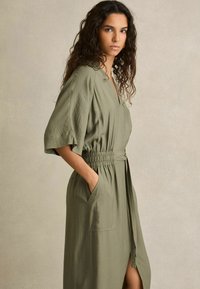 Olijfgroene jurk tot de knie met een casual pasvorm, korte brede mouwen, elastische taille en zijzakken; zachte textuur en een detail met een splits aan de voorkant.