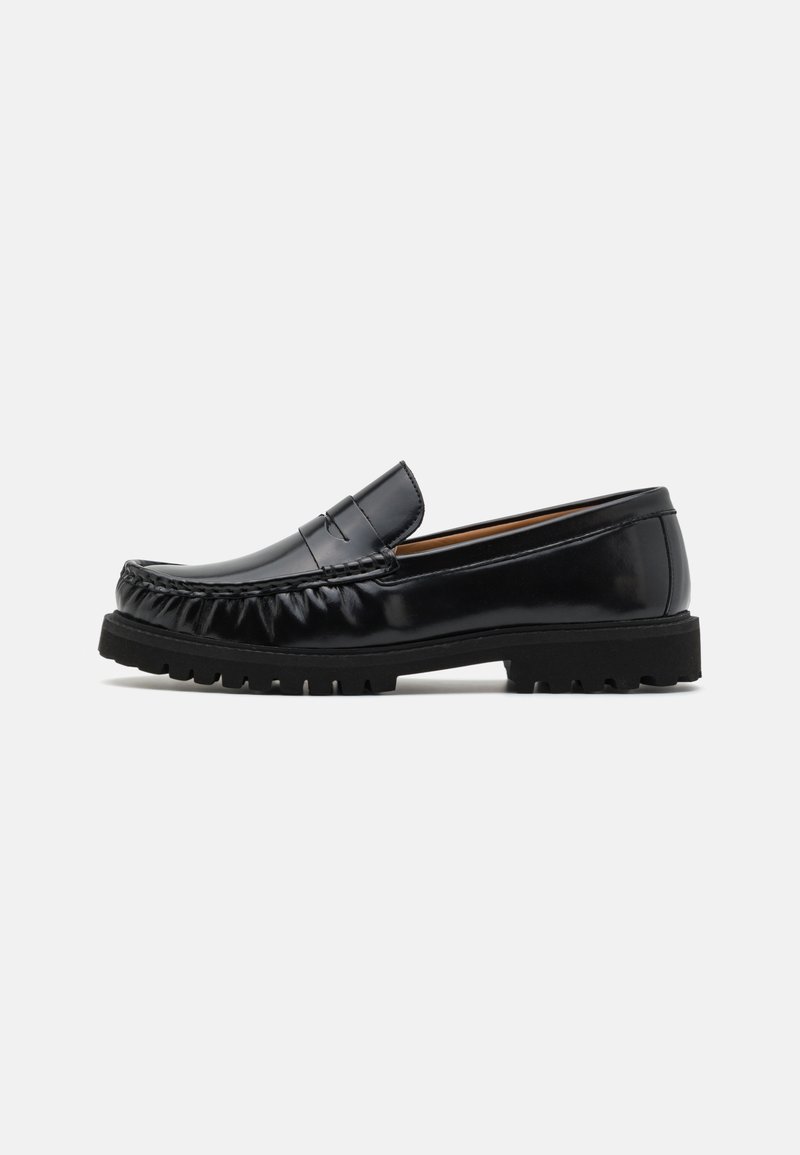 Mocassin en cuir noir avec un bout rond, couture texturée et une semelle en caoutchouc présentant des crampons proéminents pour l'adhérence et la durabilité.