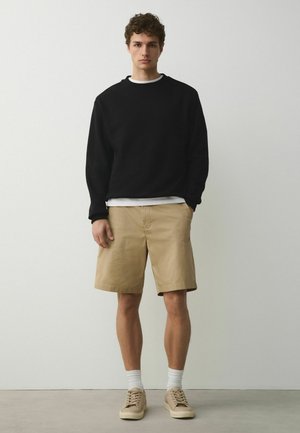 Young man standing against a plain wall wearing a black sweatshirt, beige shorts, white socks, and beige sneakers.