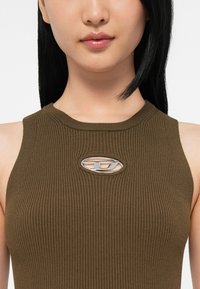 Ung person med rakt svart hår som bär en ribbad olivgrön ärmlös topp med en oval metallisk logotyputskärning vid bröstet.