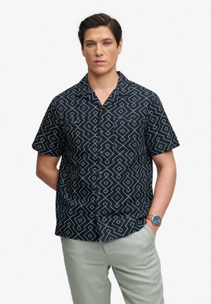 Jeune homme portant une chemise bleu marine à manches courtes avec des motifs géométriques blancs et un pantalon gris clair, debout avec une main dans la poche.