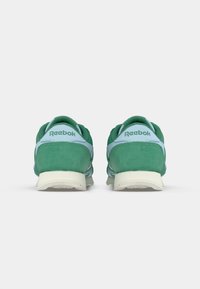 Zaļi zamšādi treniņu apavi ar gaiši zilām akcentiem, ar mīkstu zoli un Reebok zīmolu uz papēža, demonstrējot klasisku sporta dizainu.