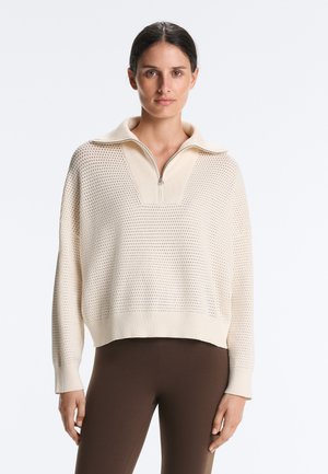 Femme portant un pull en maille crème avec un col à demi-fermeture éclair et un pantalon marron ajusté, debout devant un fond clair uni.