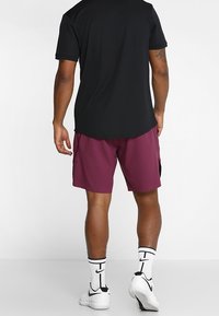 Muž stojící zády k fotoaparátu, oblečený v černém tričku s krátkým rukávem, vínových šortkách, bílých ponožkách Nike a bílých teniskách Nike.