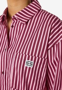 Short-sleeve shirt featuring vertical maroon and pink stripes, a button-down collar, front pocket with logo patch, and smooth fabric texture.