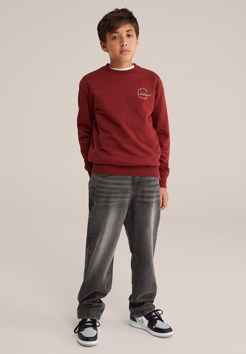 Röd sweatshirt med rund halsringning och logotypdetalj på bröstet, ihop med löst sittande mörkgrå jeans och svarta sneakers med ljusa detaljer.