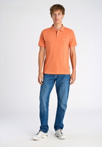 Polo arancione con due bottoni, maniche corte e logo bianco. Abbinato a jeans in denim blu e scarpe da ginnastica bianche.