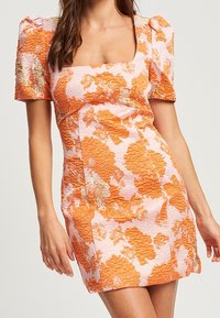 Femme portant une robe courte texturée avec un motif floral orange, des manches courtes bouffantes et un décolleté carré.