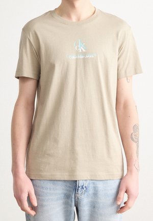 Personne portant un t-shirt beige Calvin Klein Jeans et un jean bleu clair, avec des tatouages visibles sur l'avant-bras droit.