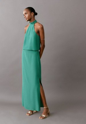 Femme portant une robe maxi verte sans manches à col montant avec une fente latérale, des sandales dorées et des bijoux en or, debout devant un fond gris.