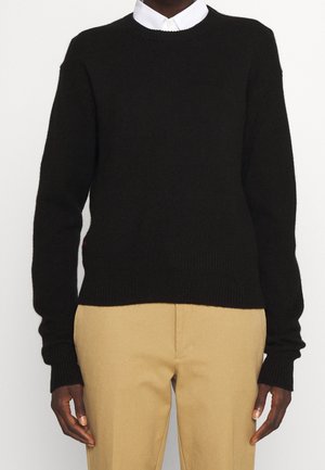 Osoba ubrana w czarny sweter crewneck na białą koszulę z kołnierzykiem i beżowe spodnie, stojąca z ramionami swobodnie opuszczonymi wzdłuż ciała.