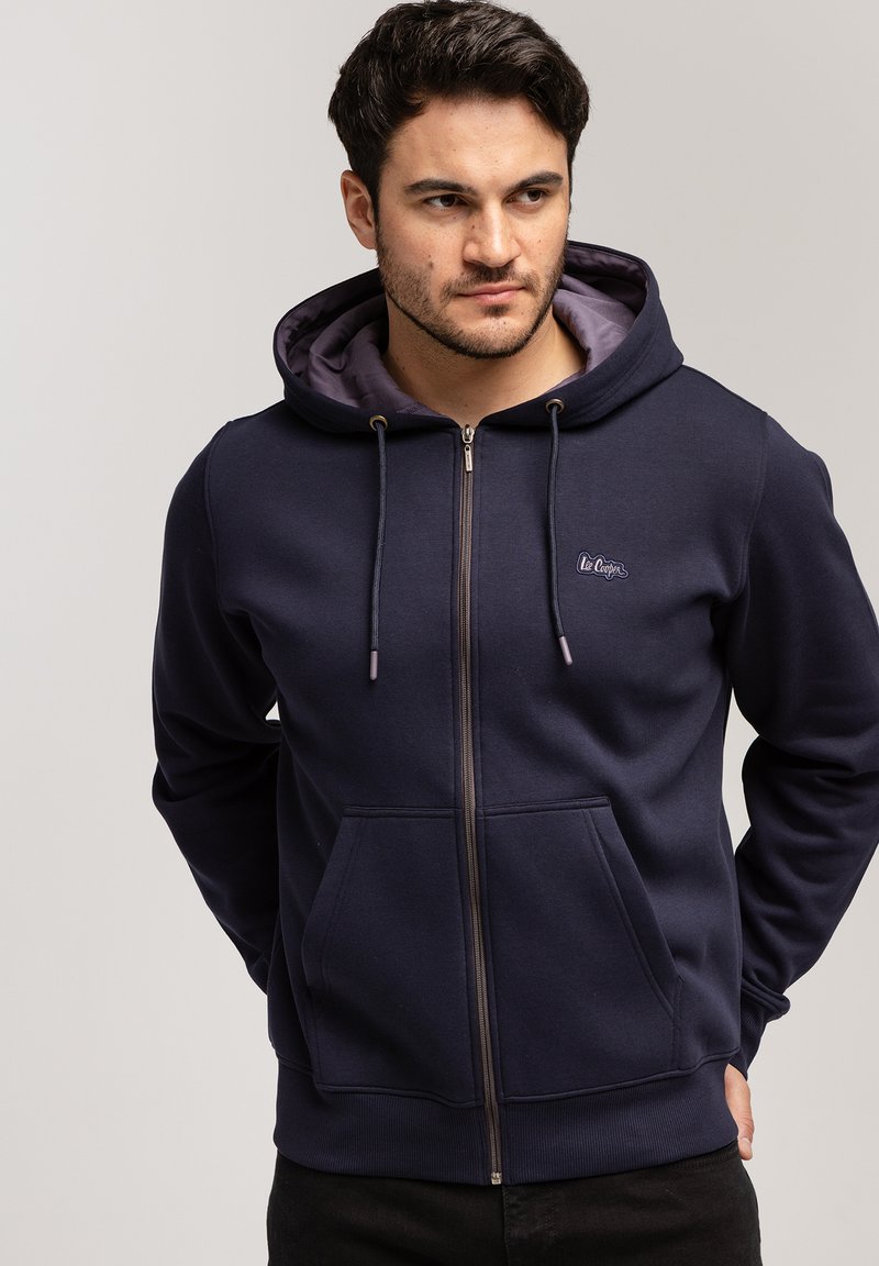 Granatowy rozpinany hoodie z kapturem na sznurku, kieszeniami z przodu i podszewką. Posiada małe logo na klatce piersiowej oraz ściągacze przy mankietach.