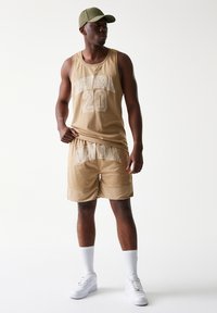 Μπεζ πλεκτό φανελάκι με τυπωμένα τα «NEW ERA» και «20» σε λευκό. Ματσάρισμα με μπεζ σορτς με παρόμοια σήμανση. Λευκά αθλητικά παπούτσια και κάλτσες, πράσινο καπέλο.