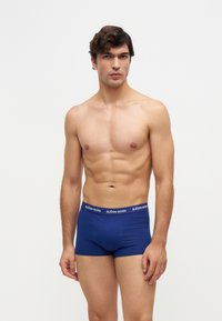 Homme musclé aux cheveux courts et bouclés portant un boxer bleu Björn Borg, debout devant un fond clair uni.