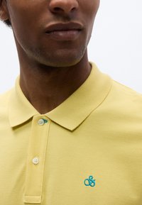 Κίτρινο πόλο μπλουζάκι με υφασμάτινη υφή, δύο κουμπιά στον γιακά και κεντημένο λογότυπο αμπερσάντ σε τυρκουάζ χρώμα στο αριστερό στήθος.