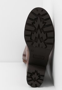 Bota de cuero marrón con suela de goma negra texturizada y estriada. Presenta un patrón en zigzag y una etiqueta de marca, colocada en ángulo sobre una superficie plana. Talla 38.