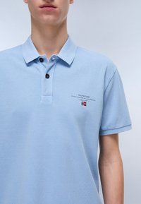 Lys blå polo skjorte med teksturert stoff, knappet krage og broderte detaljer, inkludert merkenavn og flaggaksent.